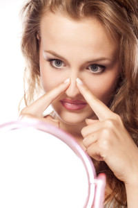 A young dark-blonde woman with her index fingers to her nose inspecting it in a mirror 